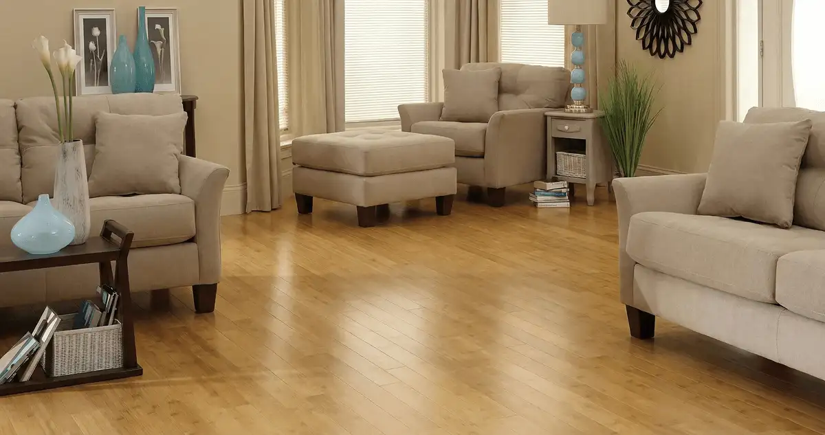 A living room with professionally installed bamboo flooring, beige sofas, a matching ottoman, a side table with a blue lamp, vases, framed pictures, and large windows with blinds and curtains in Boulder, CO
